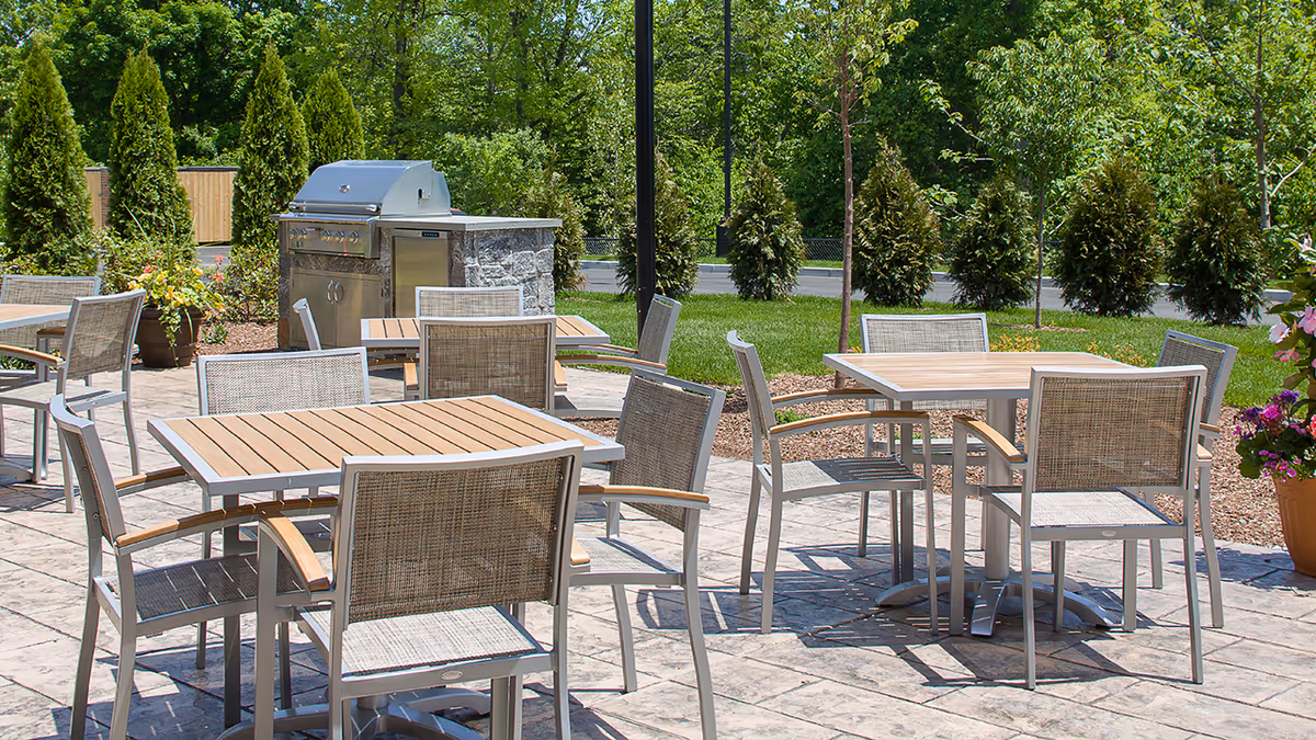 Outdoor patio area with several square wooden tables and metal-framed chairs with woven seats and backs. A stainless steel grill is visible in the background, surrounded by greenery including trees, shrubs, and potted plants.