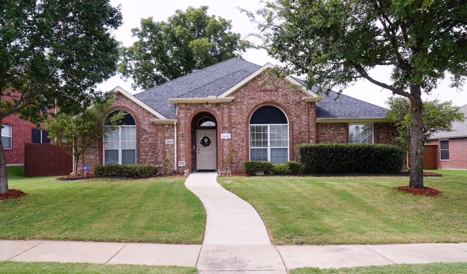 Brick senior living facility front yard with curved walkway leading to arched entrance and lush trees.