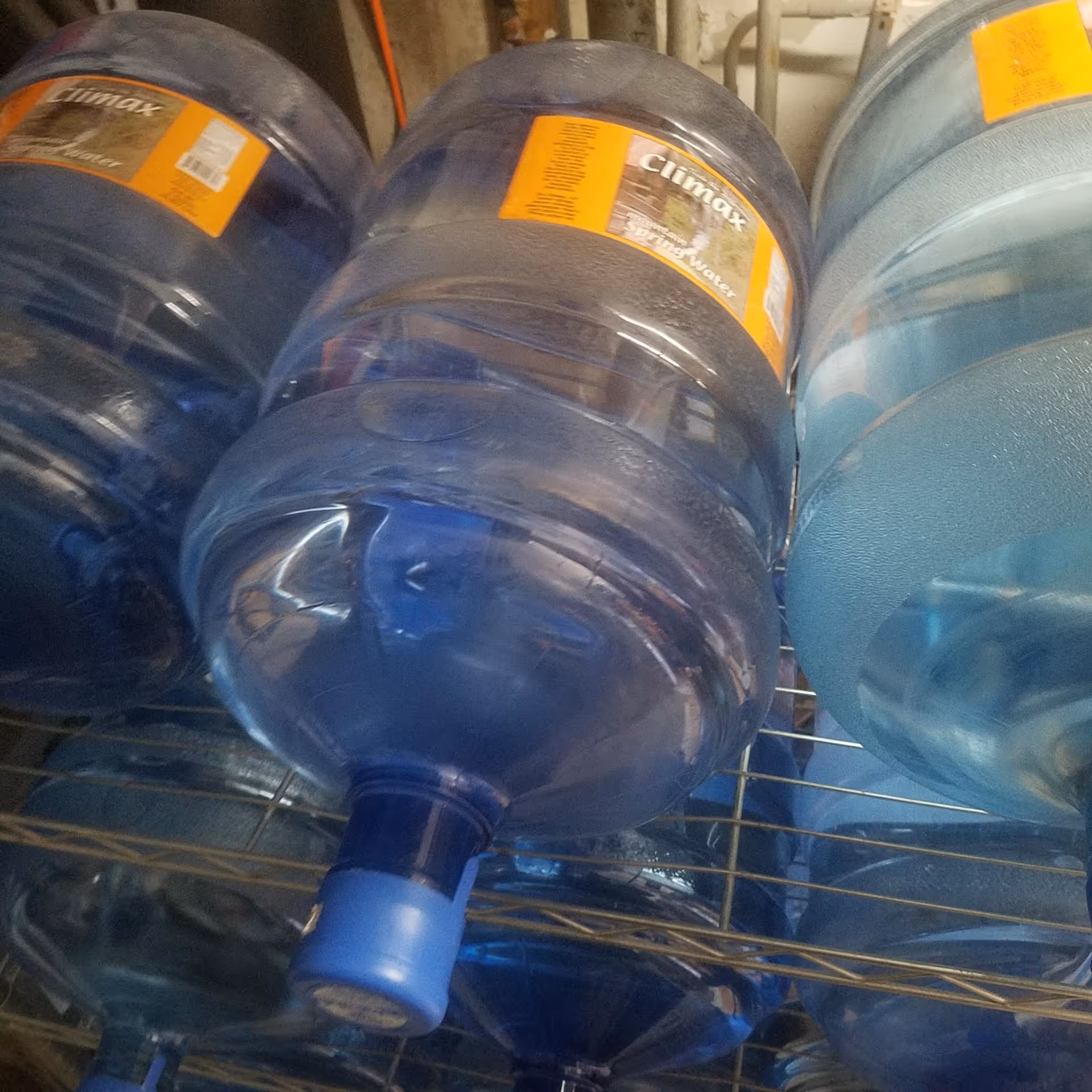 Multiple large blue water jugs stored on a metal shelf in a storage area.
