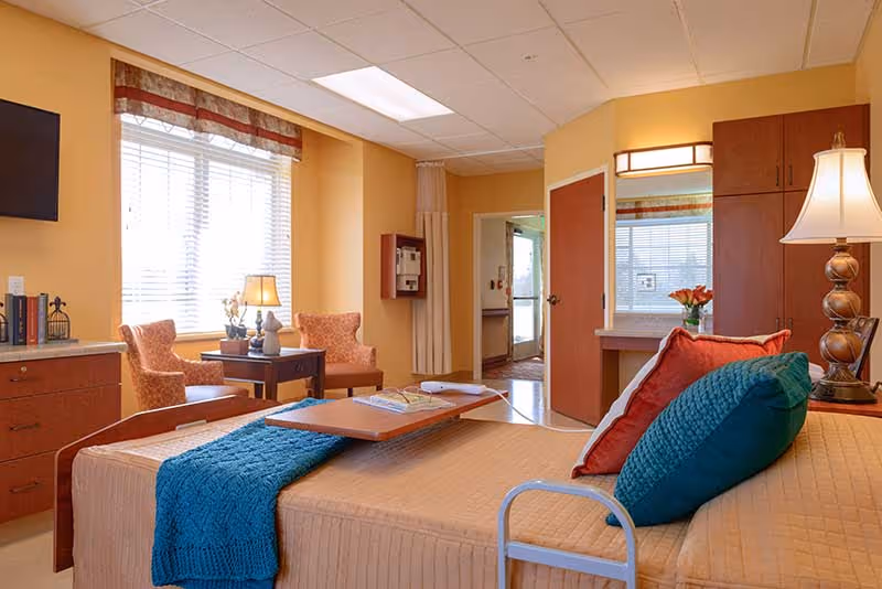 A cozy bedroom in a senior living facility with a bed featuring a beige quilt, teal and red pillows, and a teal throw blanket. There is a wooden overbed table with books on it. The room has two orange patterned armchairs next to a window with blinds and a valance. A lamp on a side table provides warm lighting. The walls are painted a soft yellow, and there is a wooden cabinet, a wall-mounted TV, and a mirror above a small desk. The door to the room is open, showing a hallway outside.