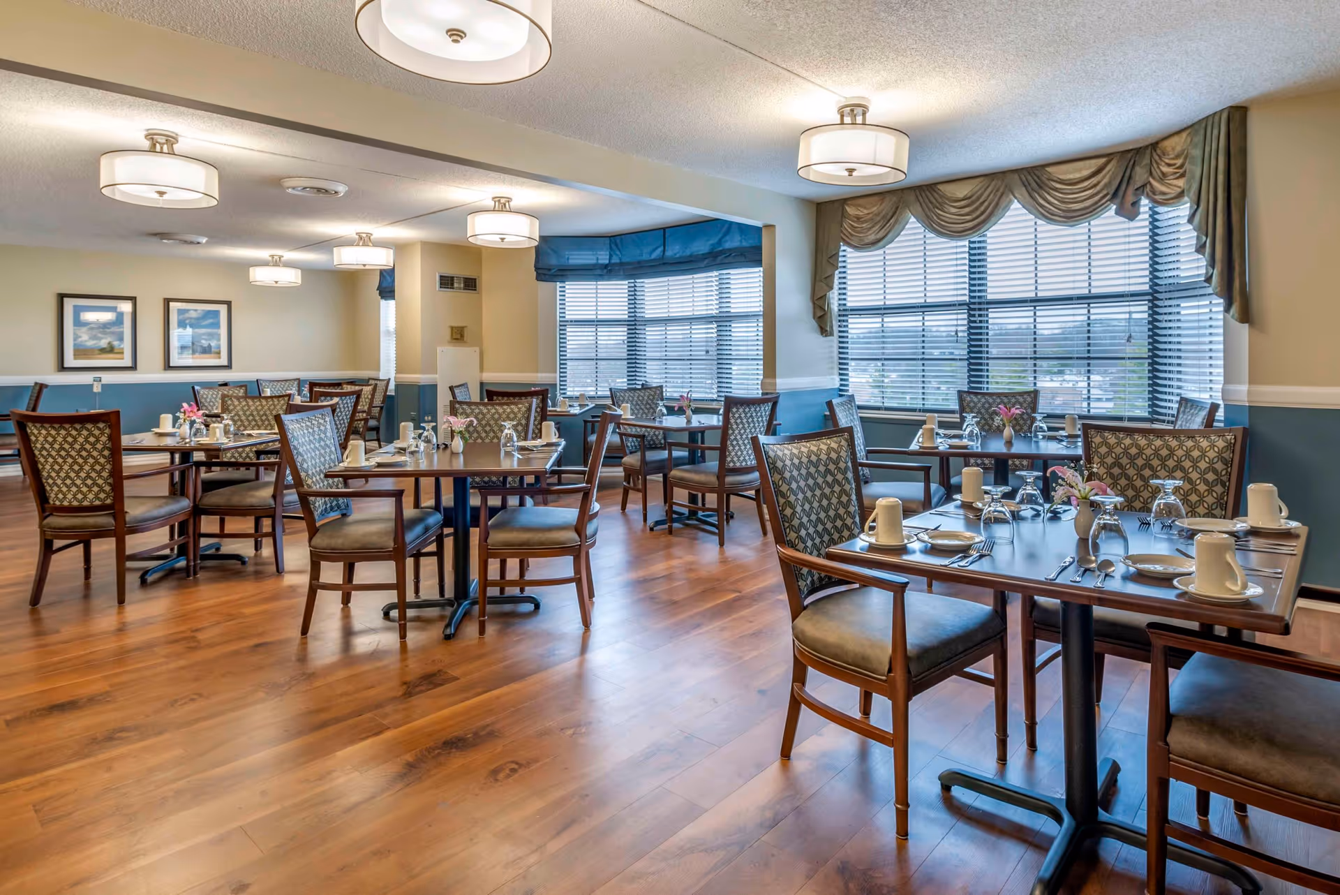 Bright dining room with multiple tables, chairs, place settings, wood floors, and large windows.