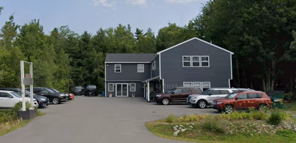 A gray two-story senior living facility with a parking lot and surrounding trees.