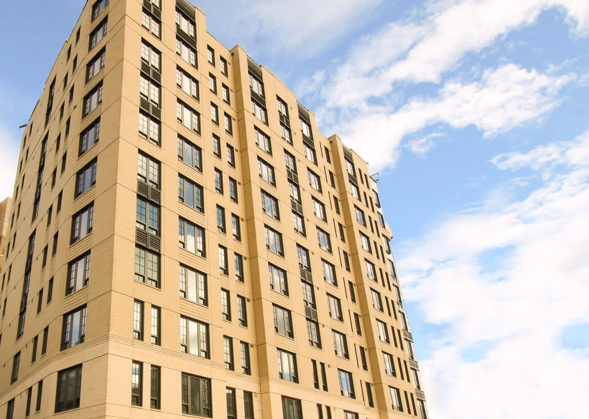 Tall beige multi-story building with many windows under a partly cloudy blue sky.