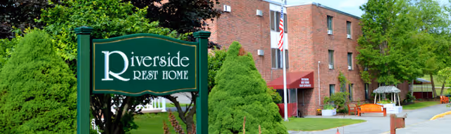 A green sign reading Riverside Rest Home stands in front of a brick nursing home building with trees, a flag, and landscaped grounds.