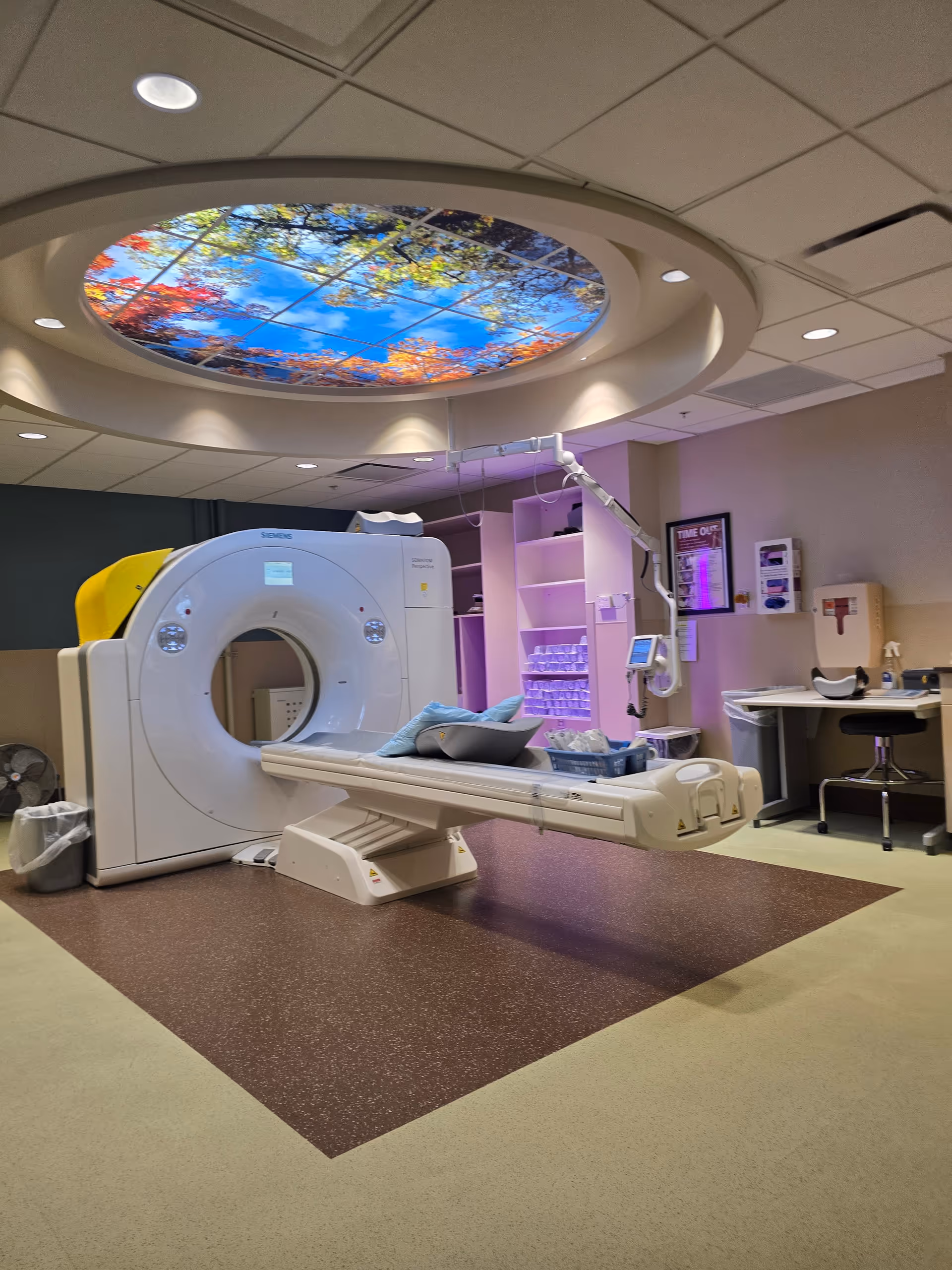 CT scanner in a hospital imaging room with a patient table and circular ceiling artwork.