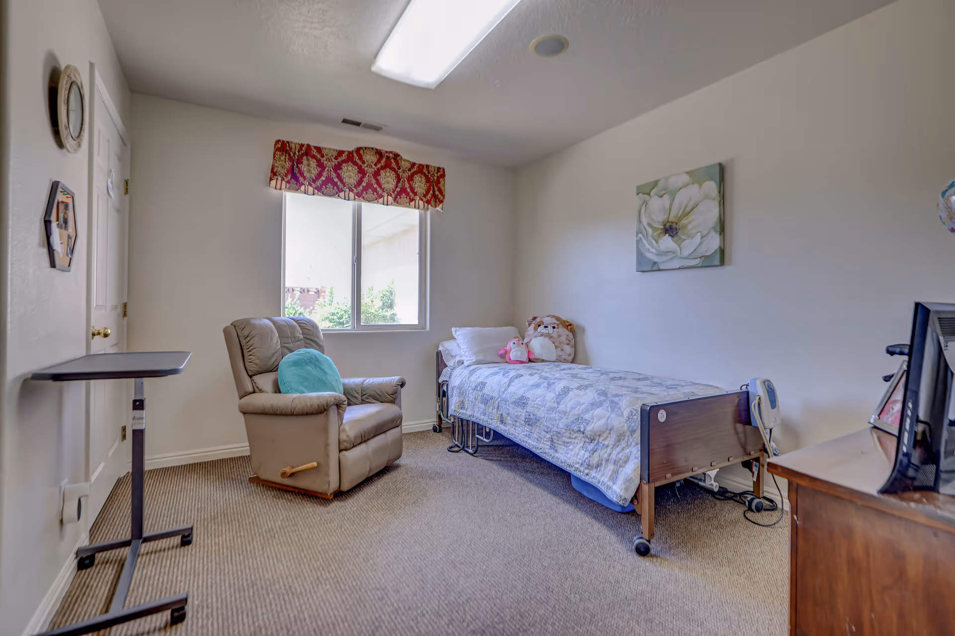A small, simple bedroom with a single bed covered in a quilt and stuffed animals on it. There is a beige recliner chair with a teal pillow next to a window with a red and gold valance. A wooden dresser with a TV on top is visible on the right side. The walls are plain white with a floral painting hanging above the bed. The room has beige carpet and a small overbed table near the door.