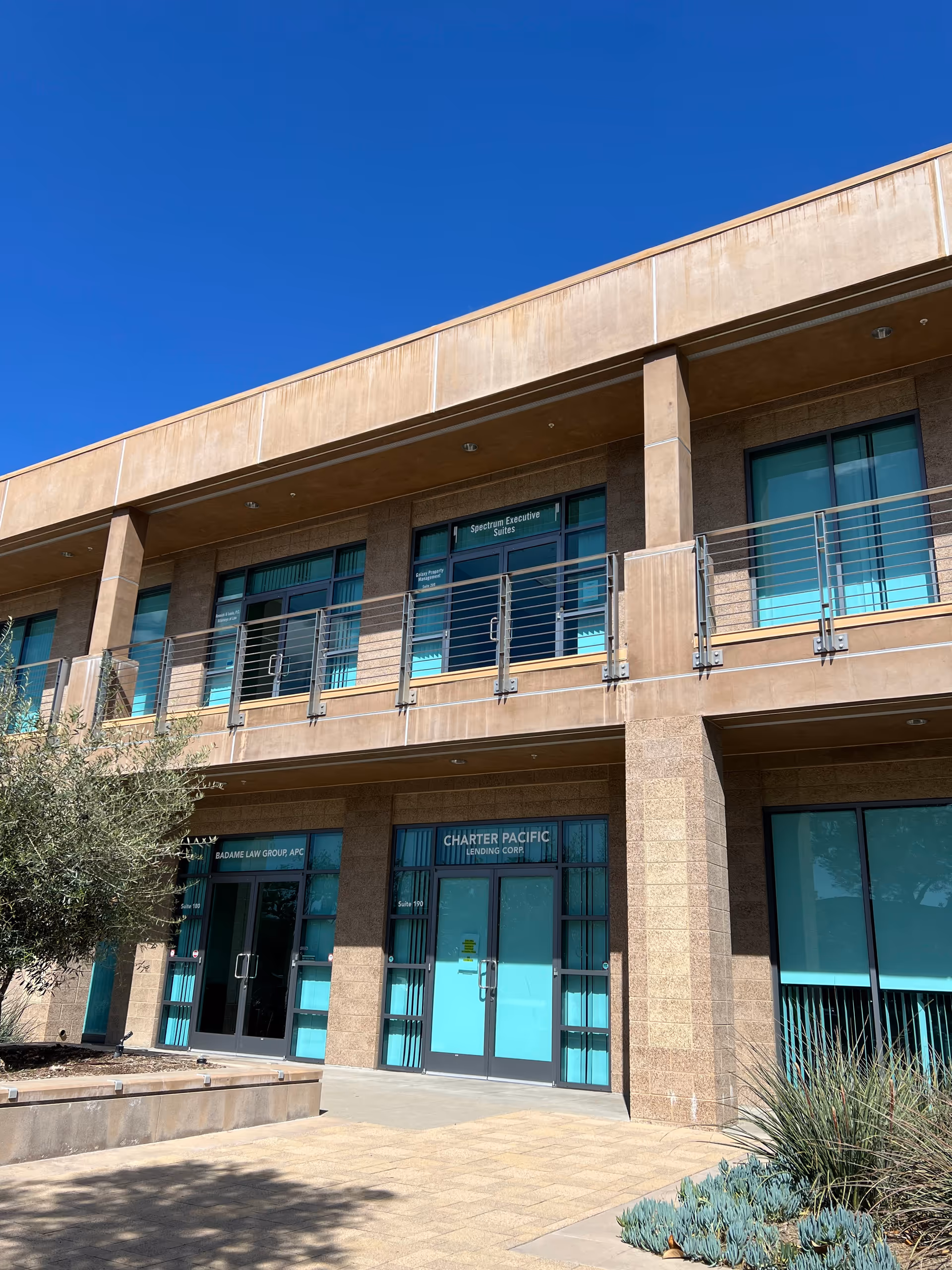 Two-story tan brick building with turquoise doors and metal railings under a clear blue sky.