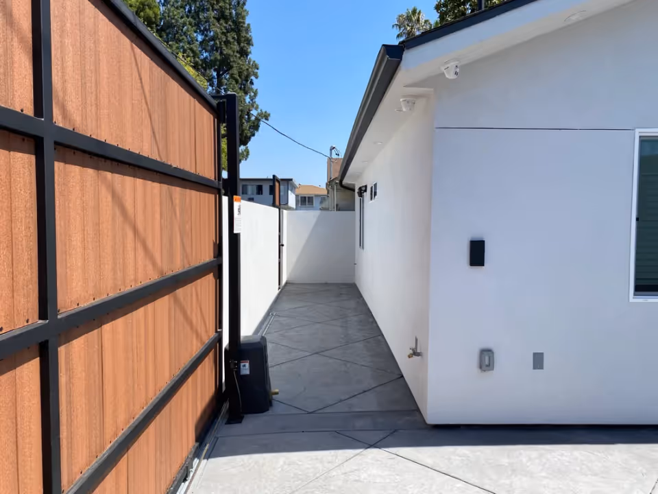 Narrow outdoor side walkway between a wood-panel gate and a white building exterior under a clear sky.