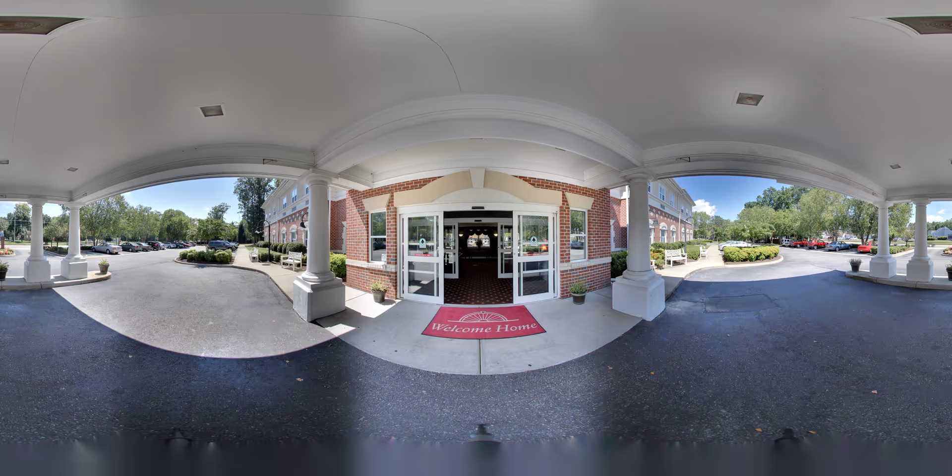 Entrance of a senior living facility with a covered driveway supported by white columns. The building has red brick walls and glass double doors with a red welcome mat that says 'Welcome Home'. There are parked cars and greenery visible on both sides of the entrance under a clear blue sky.