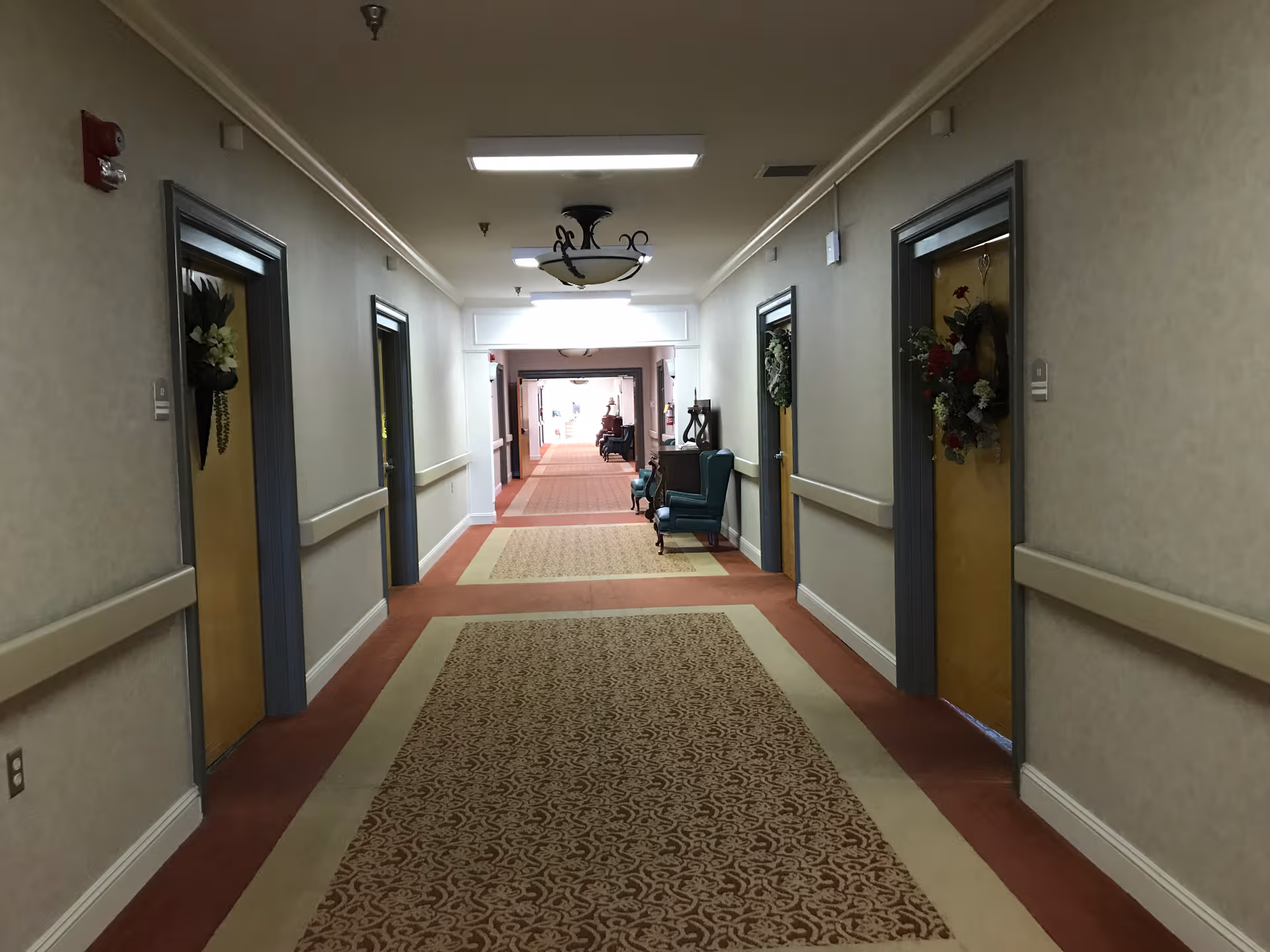 Long carpeted hallway in a senior living facility with doors and seating along walls.