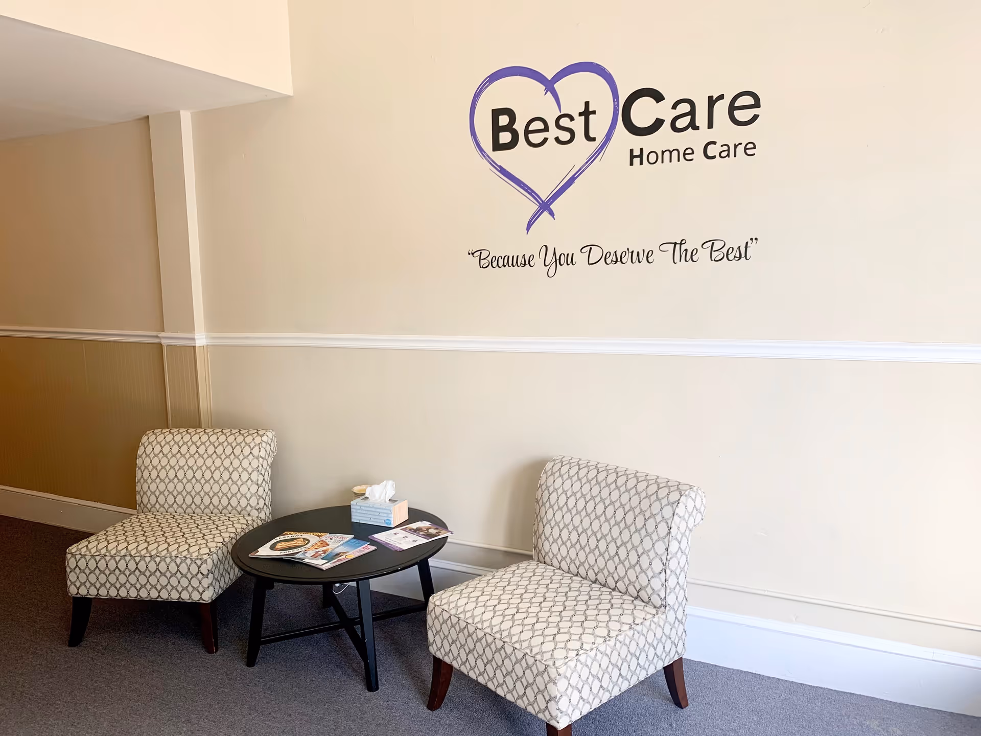 Lobby seating area with two patterned chairs and a round table beneath a Best Care logo.