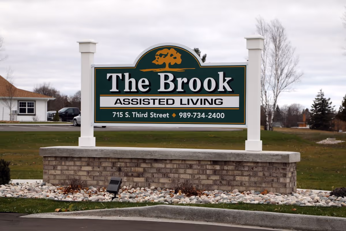 Roadside sign for 'The Brook Assisted Living' with address and phone number mounted on a brick base in front of a lawn.