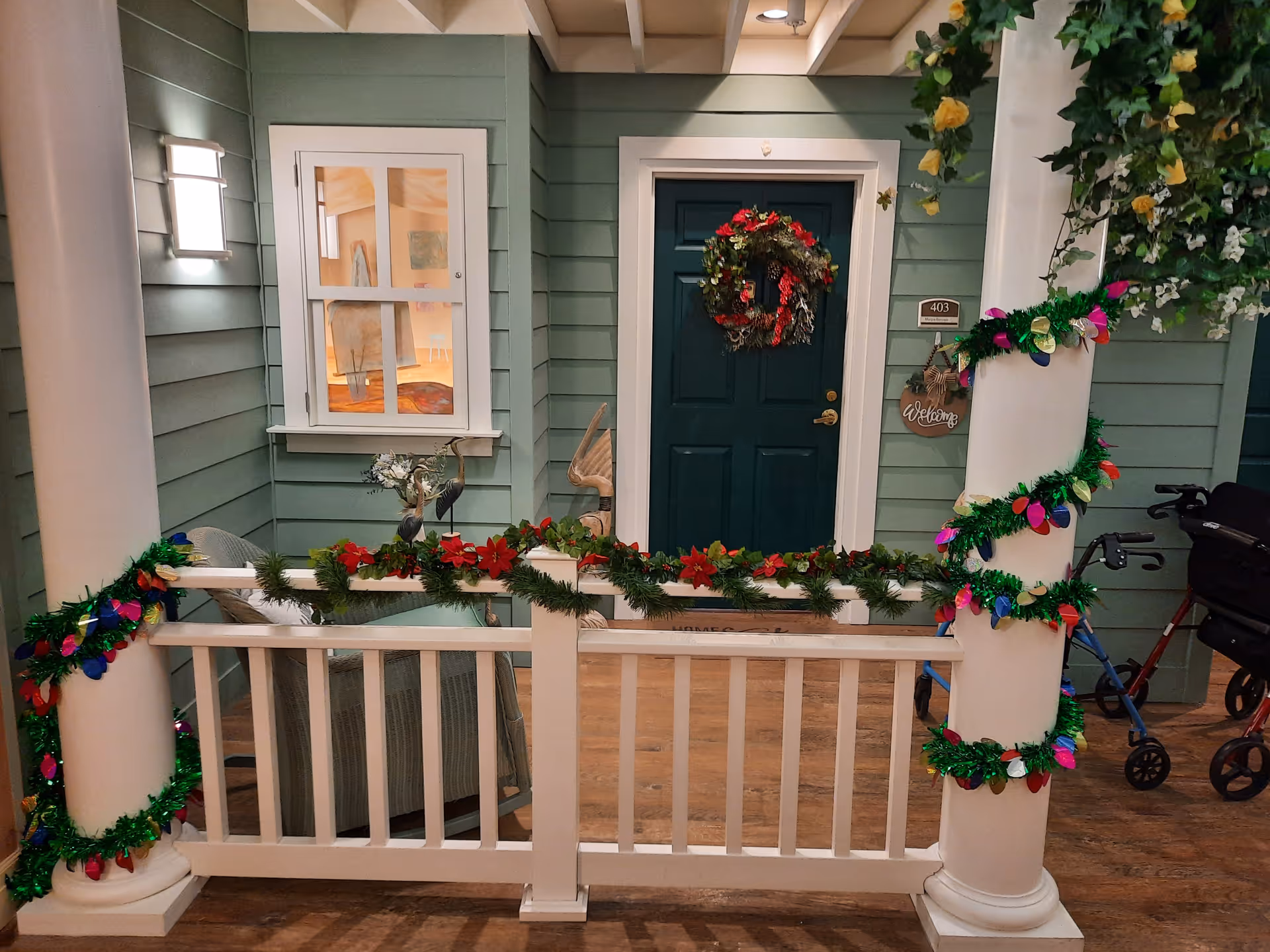 Decorated front porch of a green senior living facility with garlands, wreath, and a walker nearby.