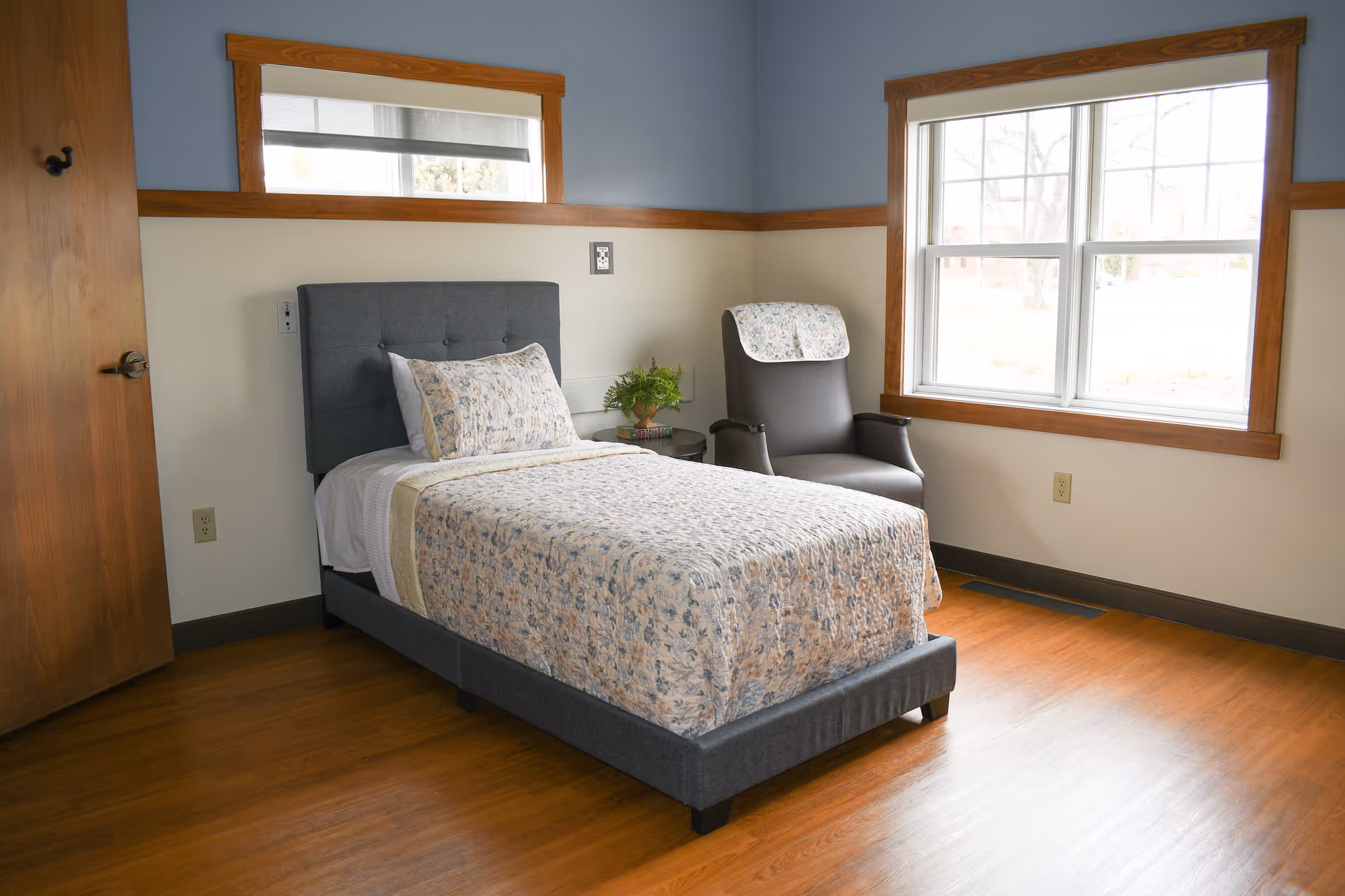 A furnished resident bedroom with a single bed, chair, window, and wooden floors.