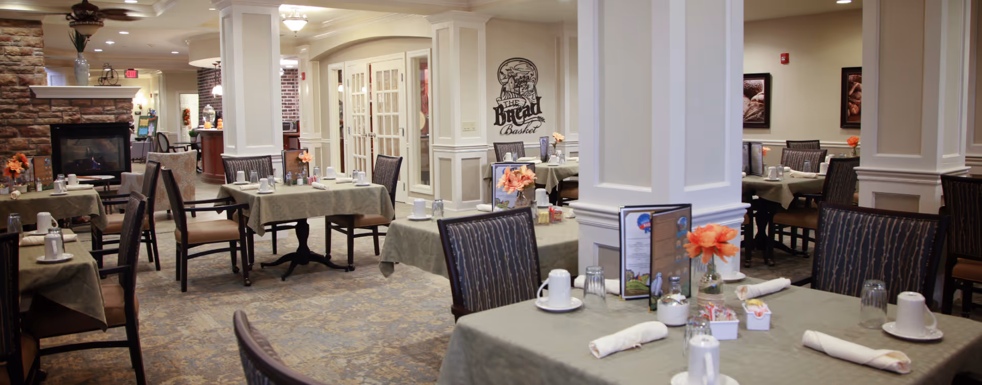 A cozy dining room in a senior living facility with multiple tables covered in green tablecloths, each set with white cups, glasses, napkins, and small flower vases. The room features a stone fireplace on the left, cushioned chairs, and soft lighting. A sign on the wall reads 'The Bread Basket.'