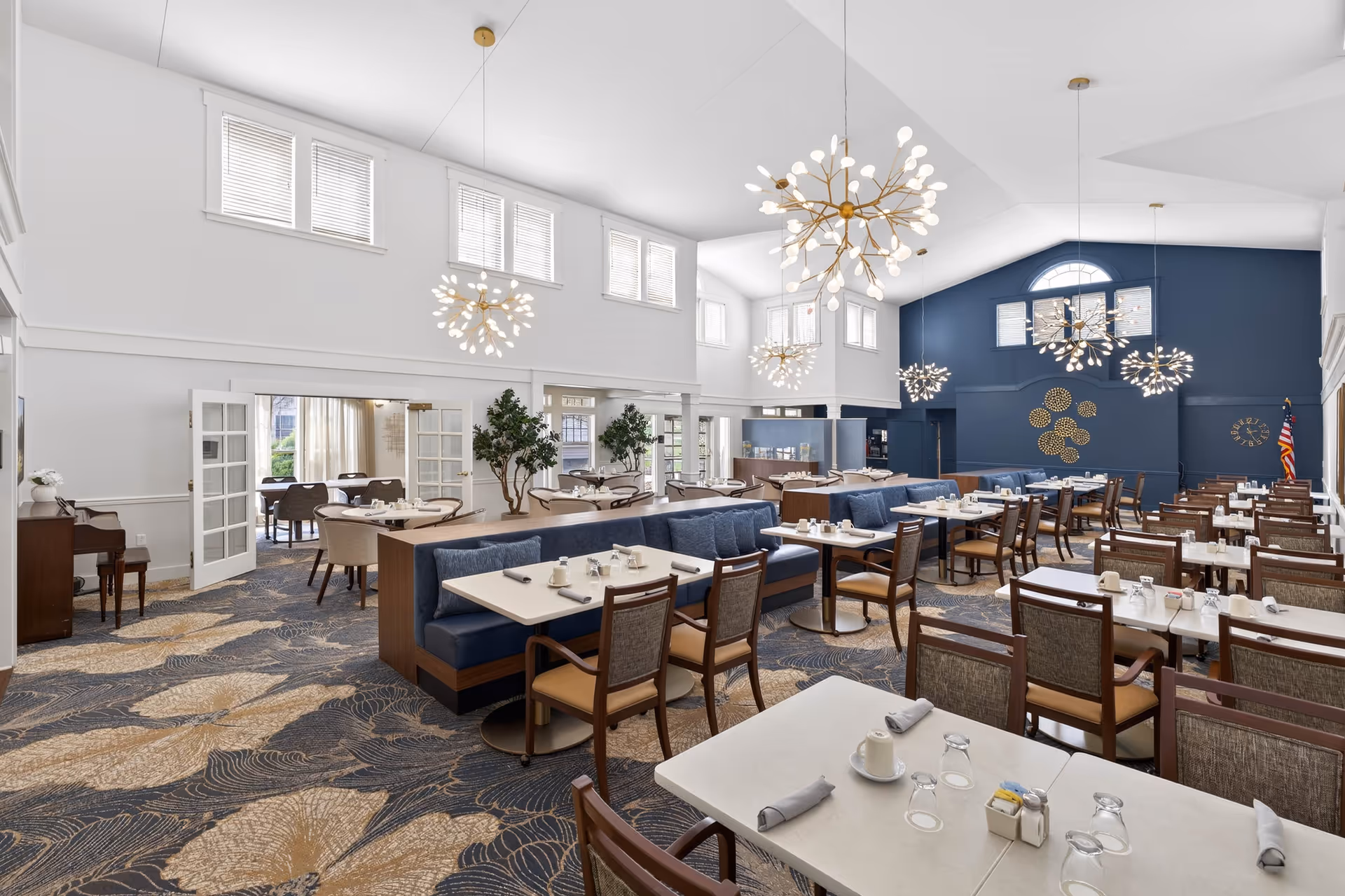 Bright, spacious dining hall with multiple tables, booths, and modern chandeliers in a senior living facility.