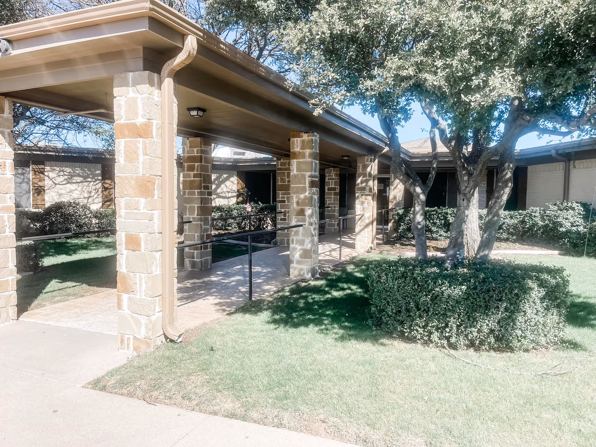 Outdoor view of a covered walkway supported by stone pillars leading to the entrance of a building, surrounded by green grass, bushes, and trees under a clear blue sky.