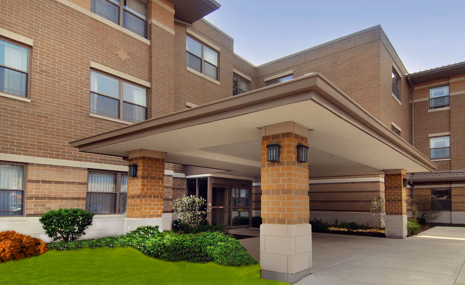 Entrance of a multi-story brick building with a covered driveway supported by brick columns, surrounded by green shrubs and small trees under a clear sky.