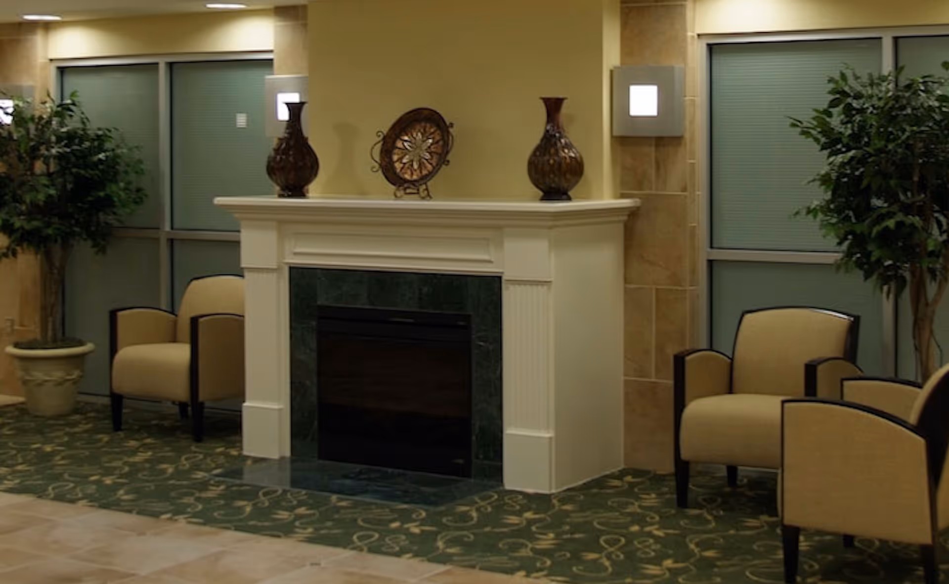 Seating area with a decorative fireplace flanked by four armchairs and potted plants in a lobby-style room.
