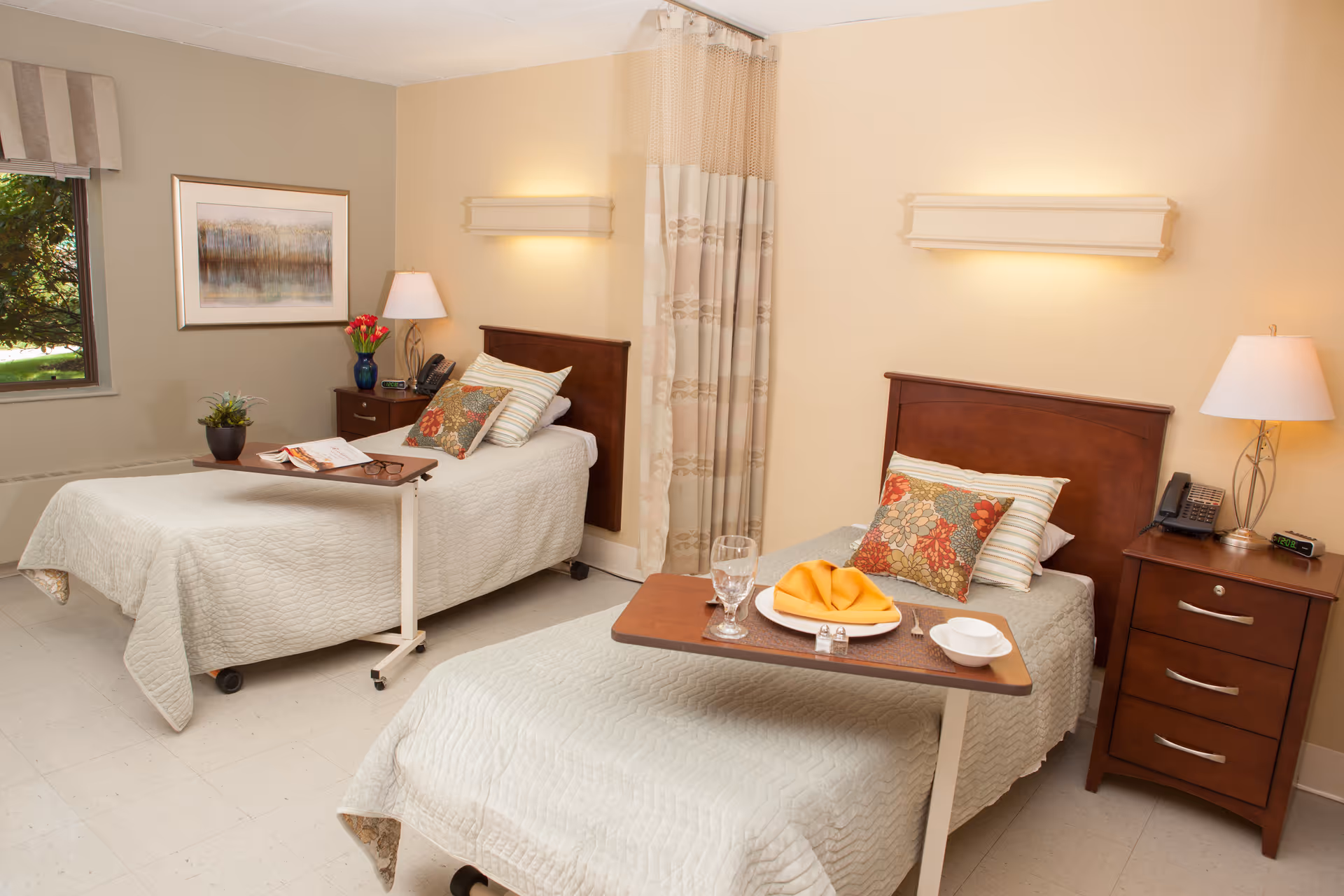 A senior living facility bedroom with two single beds, each with a wooden headboard, beige bedspreads, and decorative pillows. Each bed has an overbed table, one set with a plate, napkin, glass, and bowl. There are two wooden nightstands with lamps, a phone, and an alarm clock. A window with a valance and a framed landscape painting are on the left wall. A privacy curtain hangs between the beds.