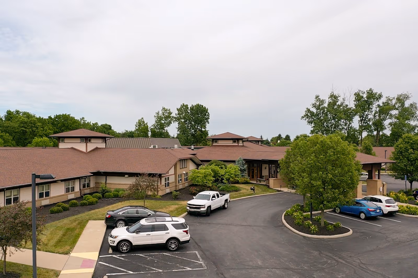Senior living campus with brown-roofed buildings, a circular driveway, and parked cars.