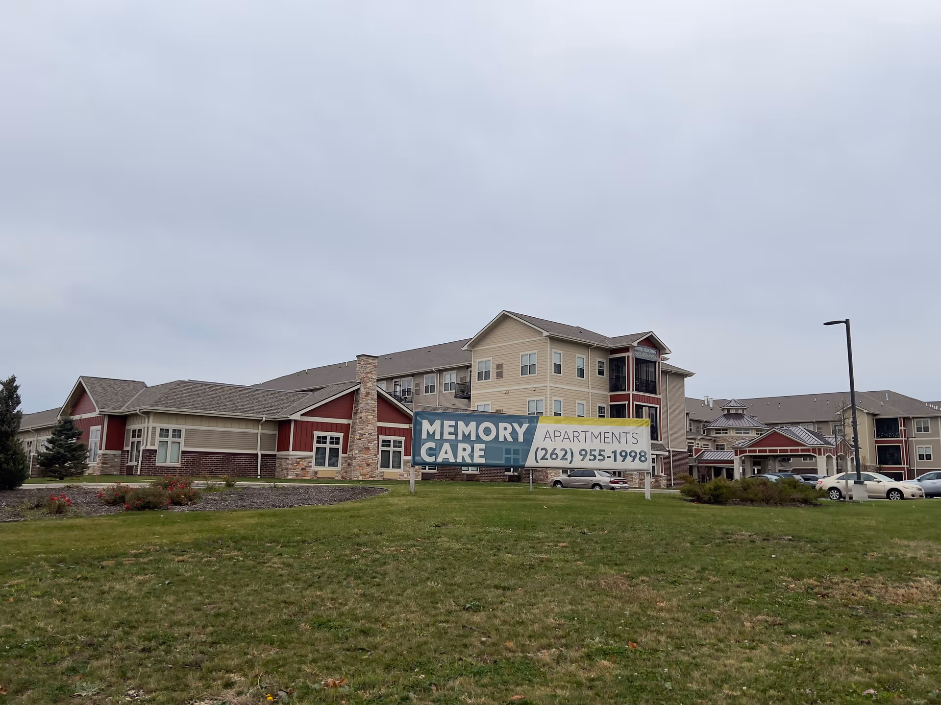 Exterior view of a multi-story memory care apartment building with a large sign on the front lawn reading "MEMORY CARE APARTMENTS (262) 955-1998".