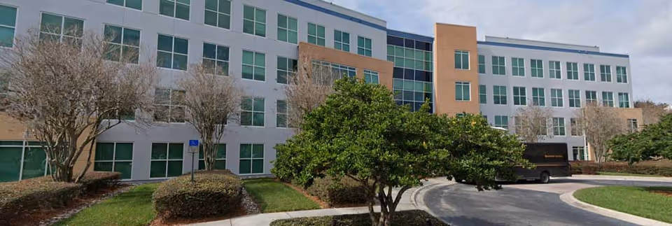 A modern multi-story senior living facility with a curved road, trees, and a parked bus.