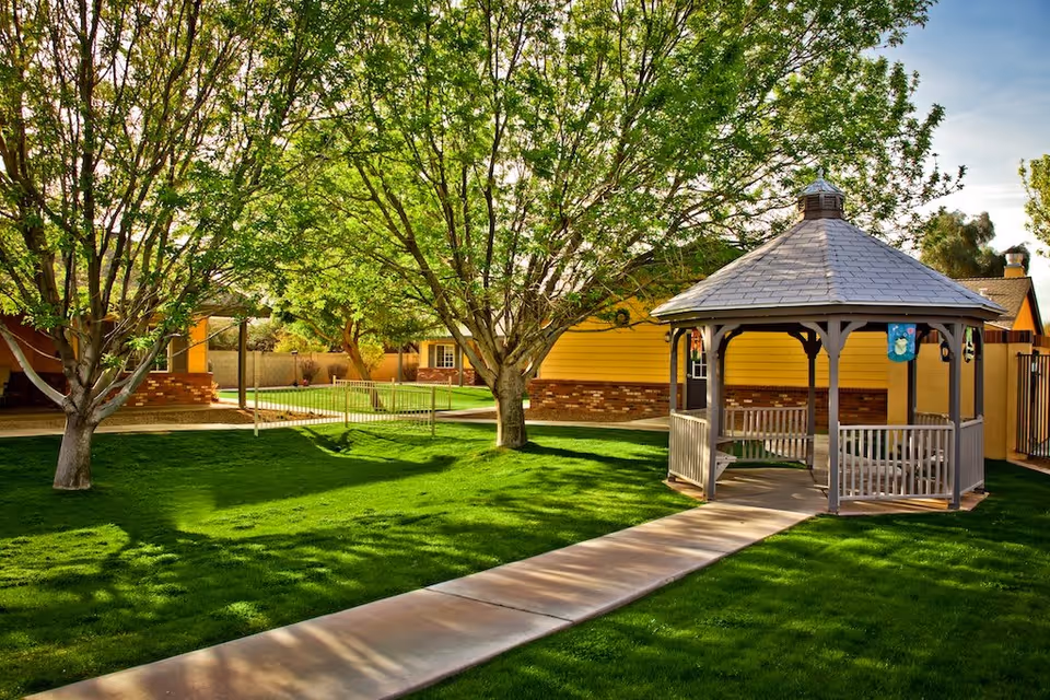 A sunny outdoor garden area with a paved walkway leading to a wooden gazebo. The gazebo has a gray shingled roof and is surrounded by green grass and leafy trees. In the background, there are yellow buildings with brick accents.
