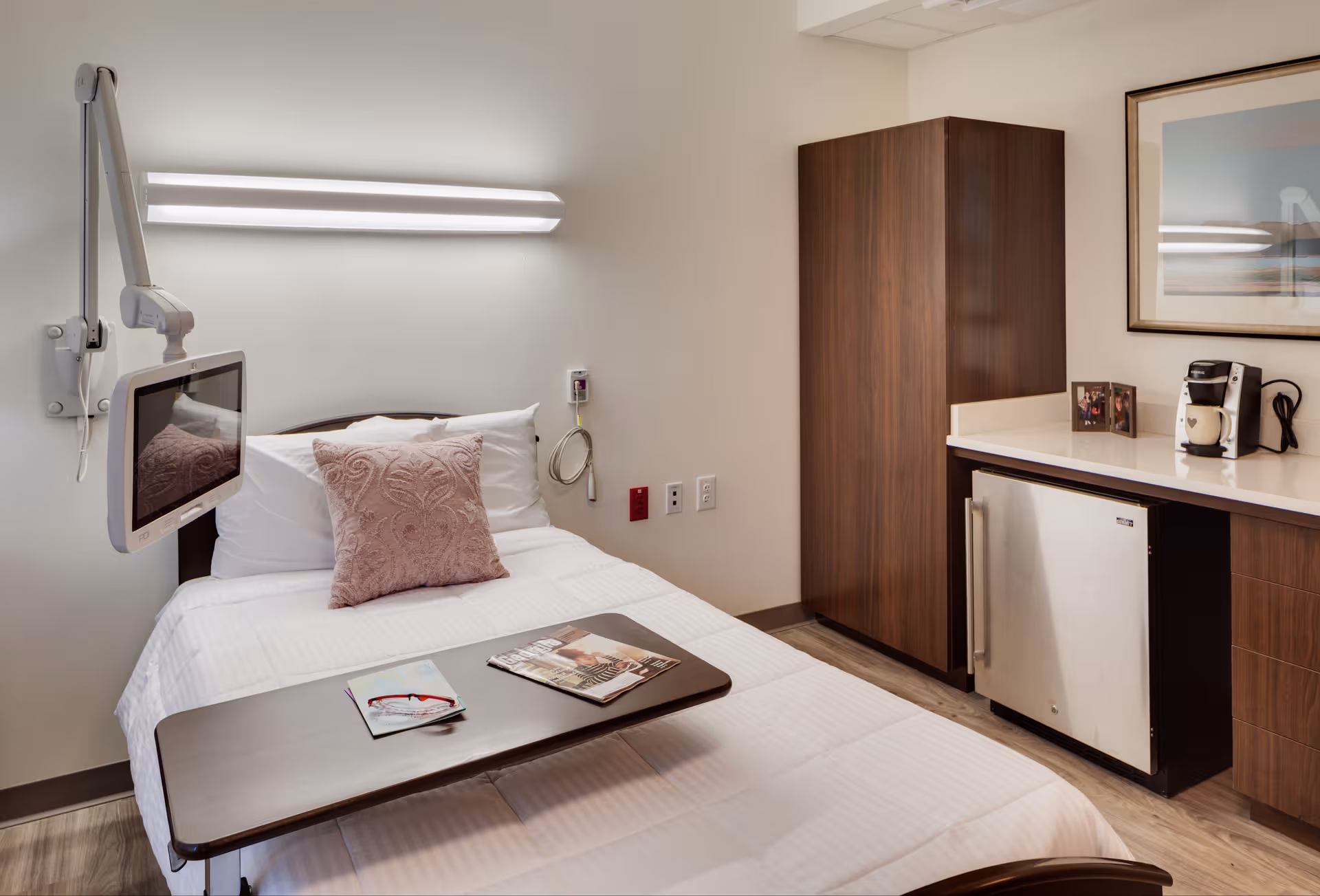 A hospital-style senior living bedroom featuring a bed, wall monitor, cabinet, and small kitchenette.