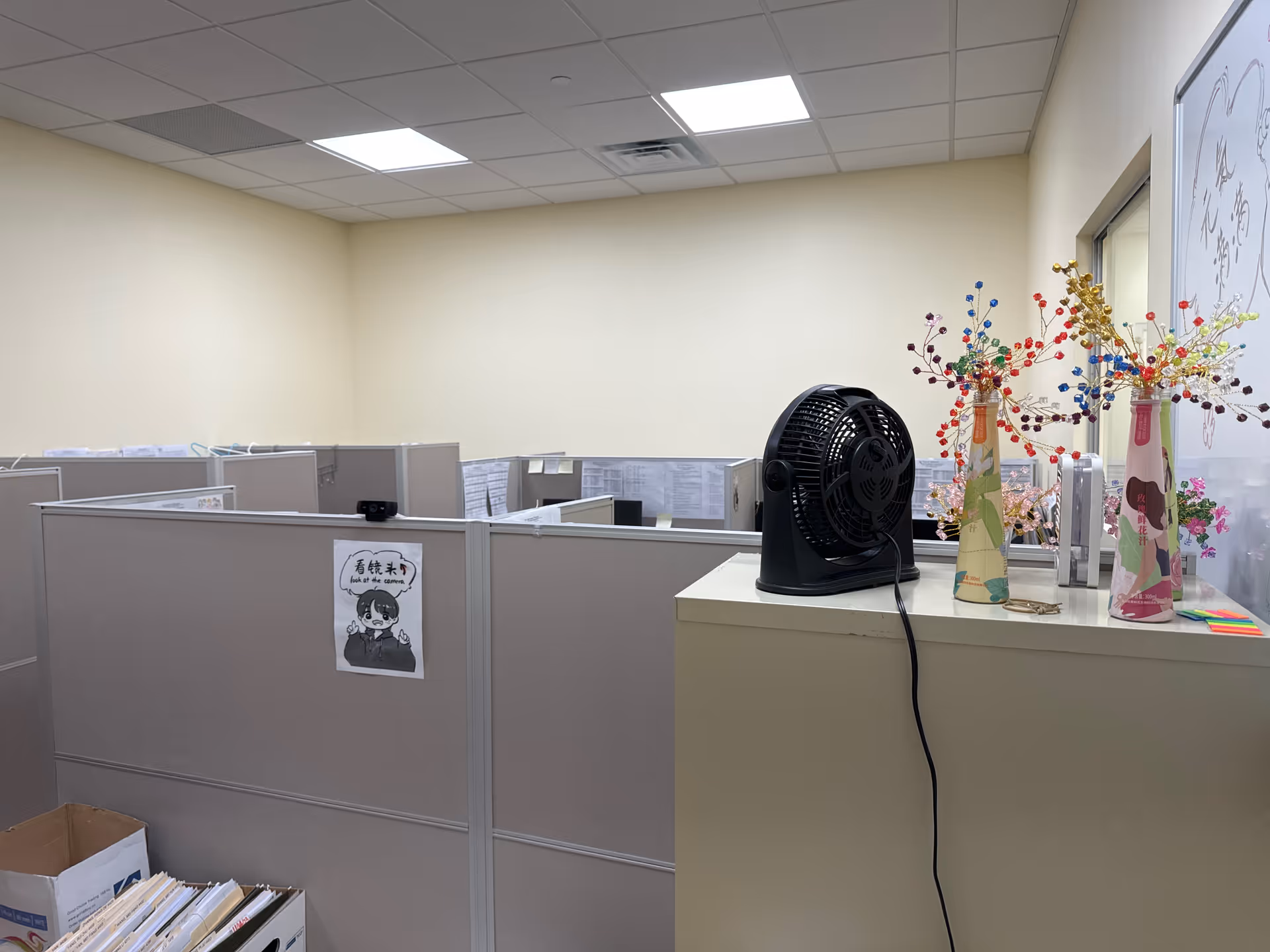 Office cubicles and a desk with a fan in a neutral-lit workspace.