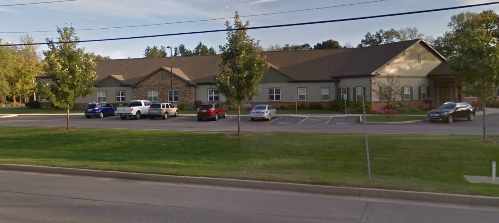 Front exterior of a one-story senior living facility with a lawn, parking lot, and several parked cars.