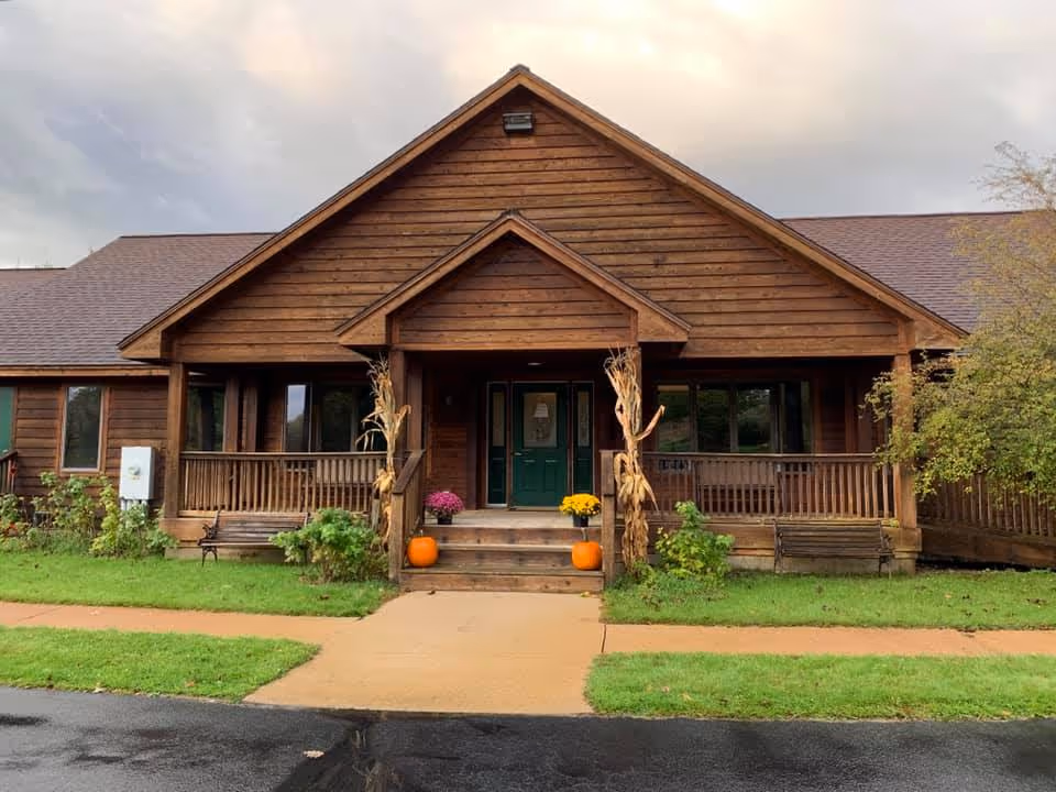 Front exterior of a wooden single-story building with a covered porch, green double doors and autumn decorations including pumpkins on the steps.