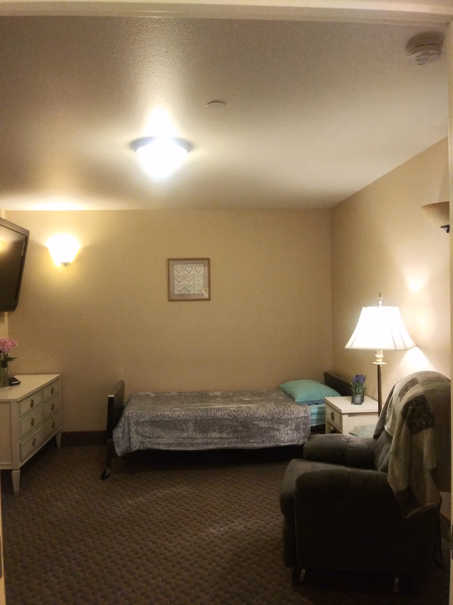 A simple, well-lit bedroom in a senior facility with a bed, dresser, and chair.