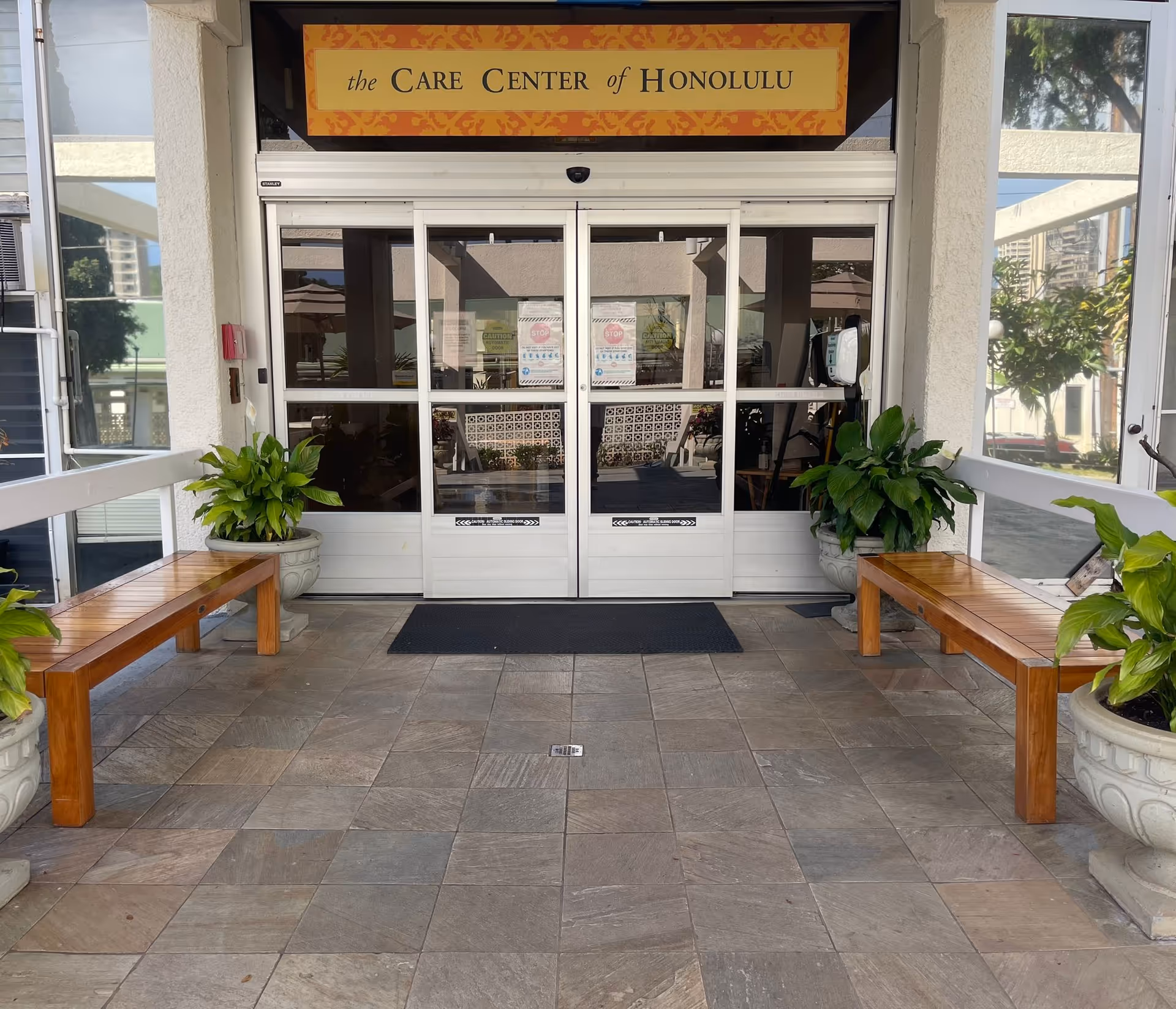 Entrance to The Care Center of Honolulu with glass doors, benches, and potted plants.