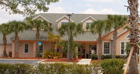 Single-story peach-colored senior living building entrance lined with palm trees and a parking area.