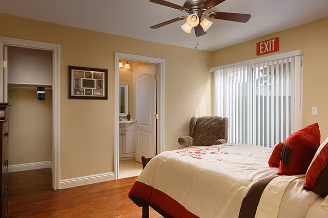 Cozy beige bedroom with a bed, red pillows, armchair, ceiling fan, and sliding glass door blinds.