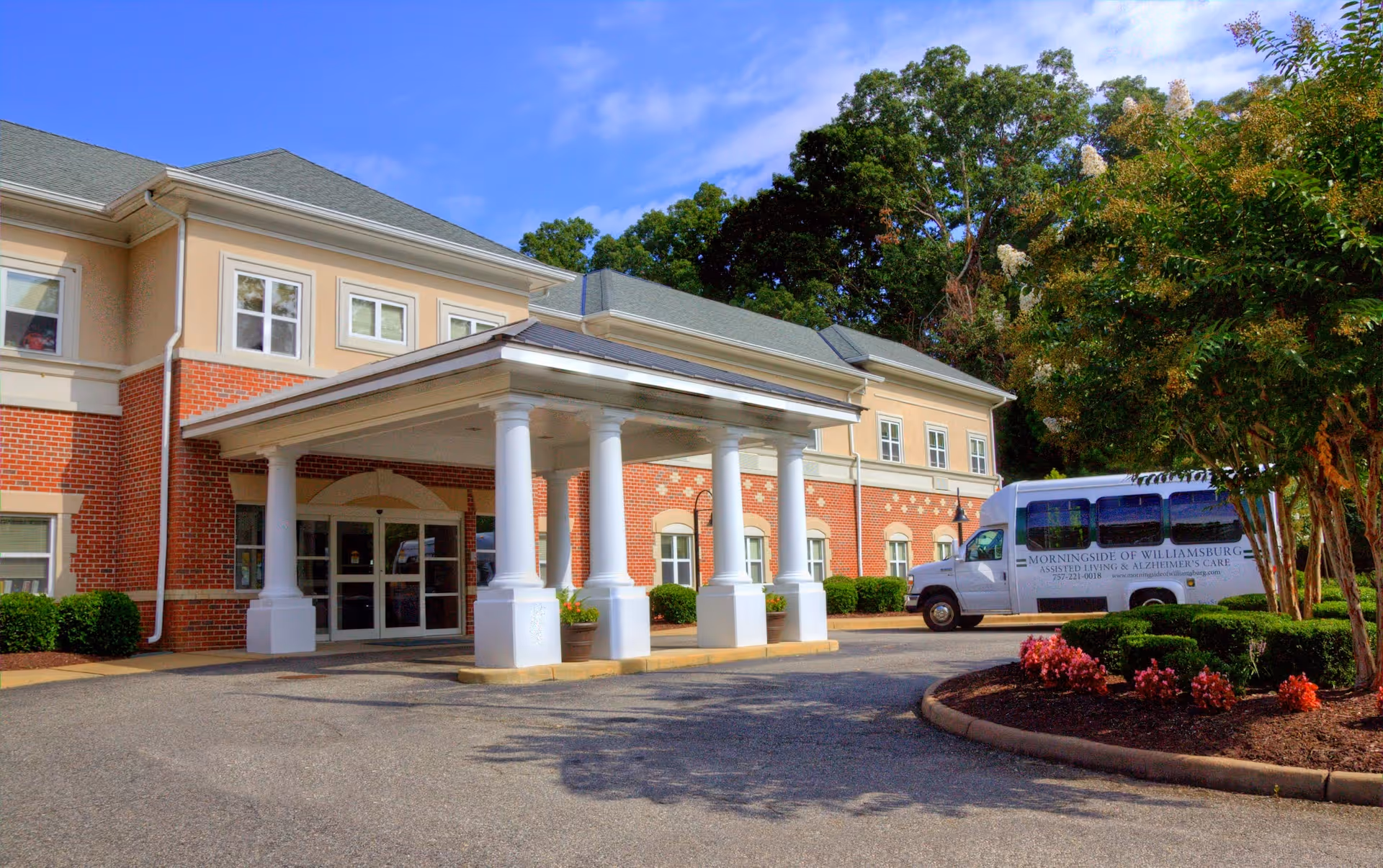 Exterior view of a senior living facility building with a covered entrance supported by white columns. There is a driveway in front and a white shuttle van parked to the right. The building has red brick and beige siding with multiple windows. Trees and landscaping with flowers are visible on the right side under a blue sky with some clouds.
