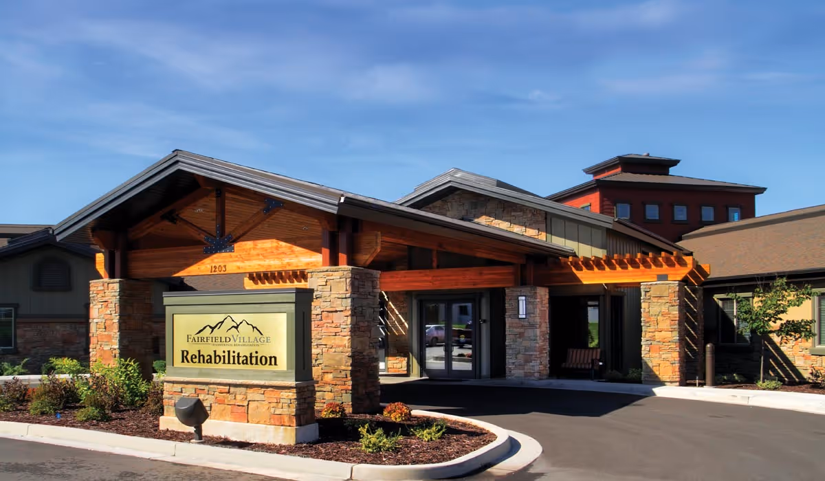 Front entrance of the Fairfield Village Rehabilitation building with stone pillars, a wooden canopy, and a landscaped driveway.
