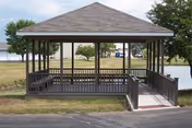 An outdoor gazebo with railings sits on a grassy area near a pond.
