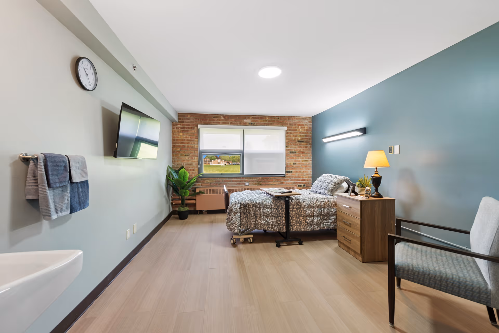 Senior living bedroom with bed, dresser, chair, TV, brick accent wall, and window.
