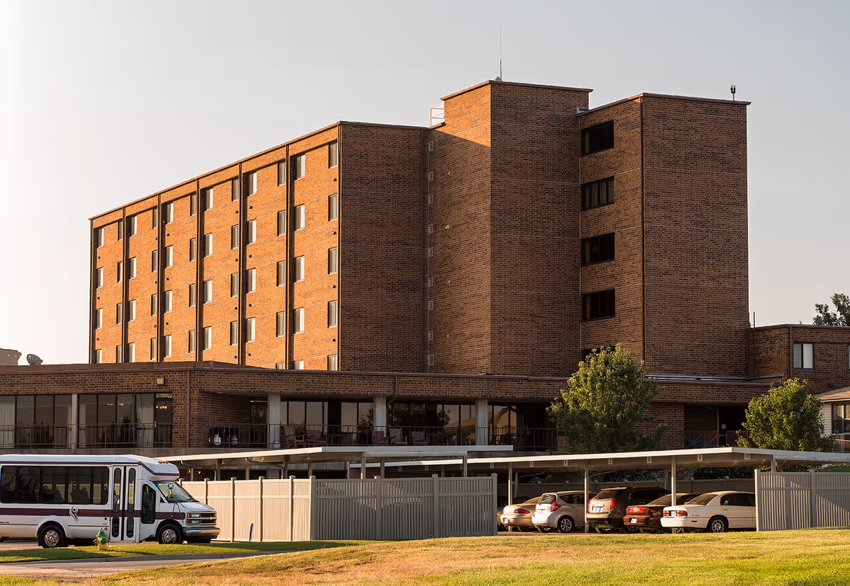 Exterior view of a multi-story brick building with several windows, a covered parking area with cars, a white shuttle bus parked nearby, and some trees and grass in the foreground under a clear sky.