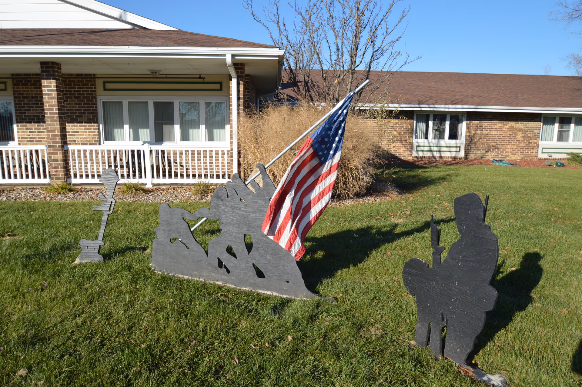 Outdoor scene showing a green lawn with black silhouette cutouts of soldiers, one holding an American flag, in front of a brick building with white railings and windows.