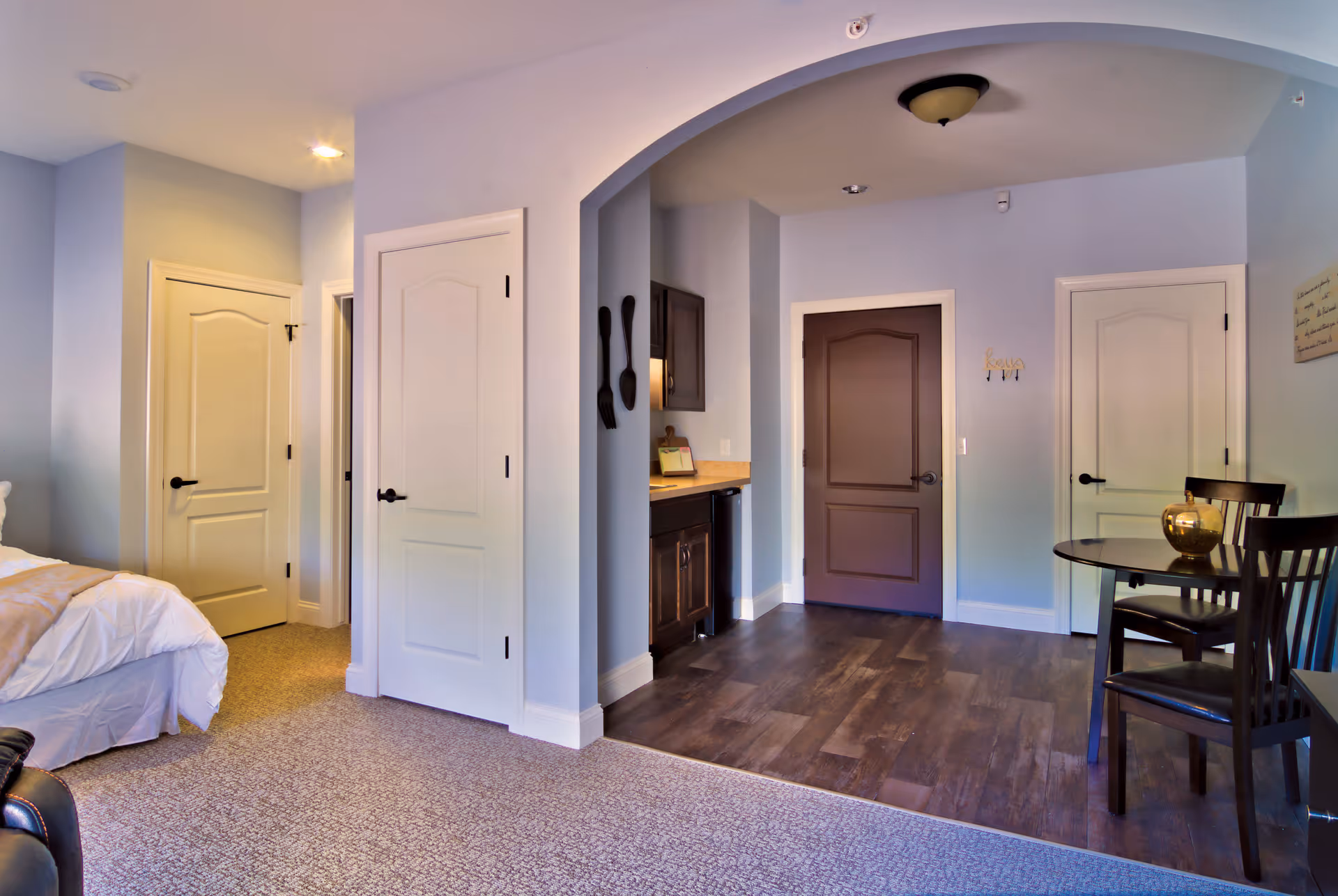 Interior view of a senior living facility room showing a bed partially on the left, two white doors, a dark brown door, a small kitchenette area with cabinets and countertop, and a small round dining table with two chairs and a decorative item on top. The flooring transitions from carpet in the bedroom area to wood in the kitchenette and dining area.
