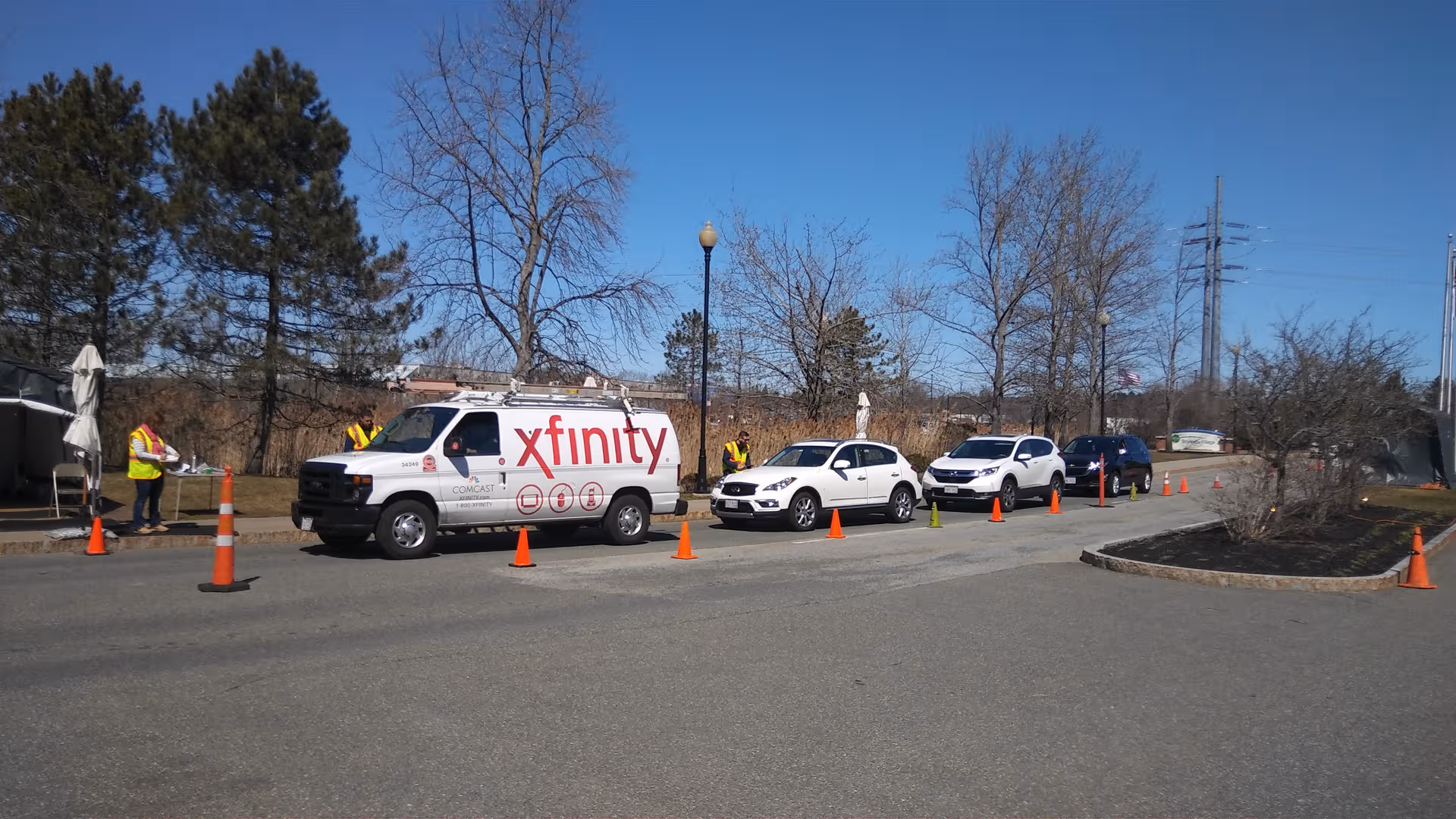 Outdoor parking lot with an Xfinity van, several cars, and workers beside orange safety cones.
