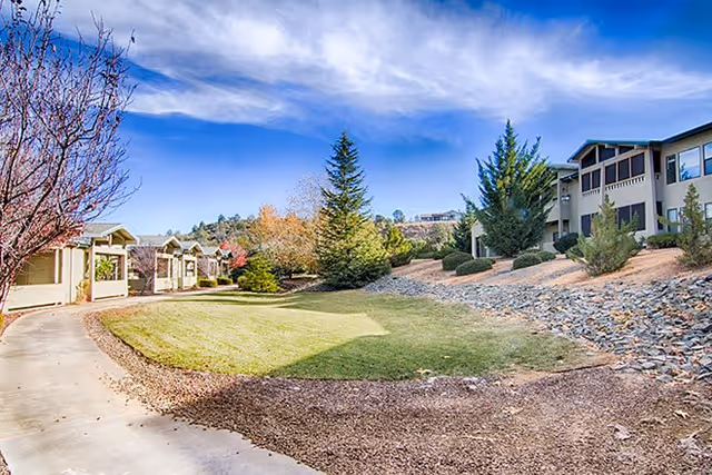 Outdoor view of Evergreen Village Prescott showing a landscaped area with a curved concrete pathway, green grass, trees, shrubs, and buildings in the background under a partly cloudy blue sky.