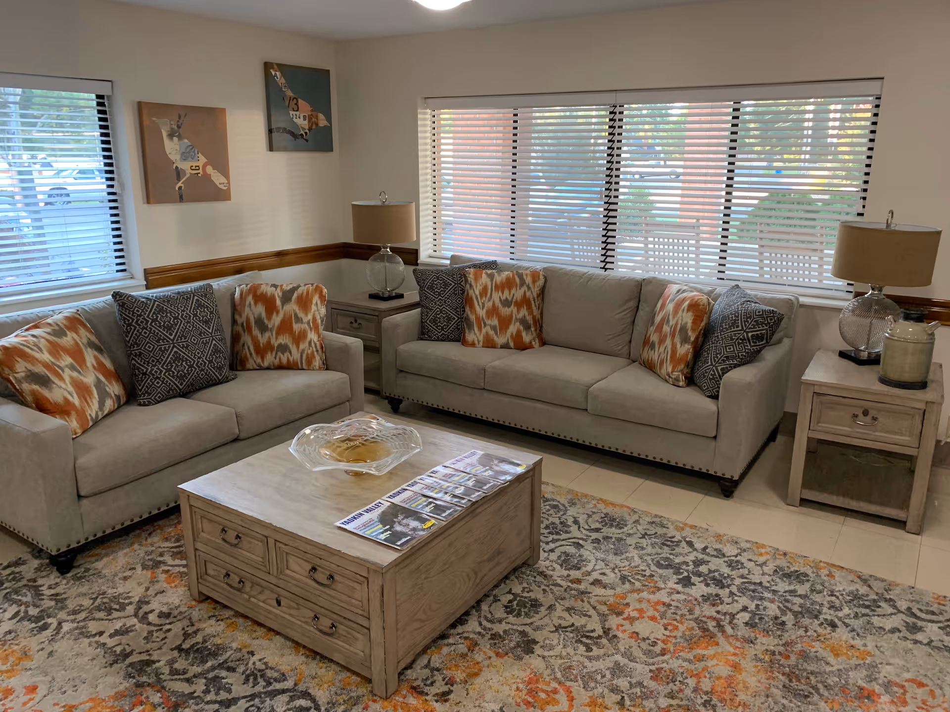 Bright senior living lounge with two beige sofas, coffee table, and large window blinds.