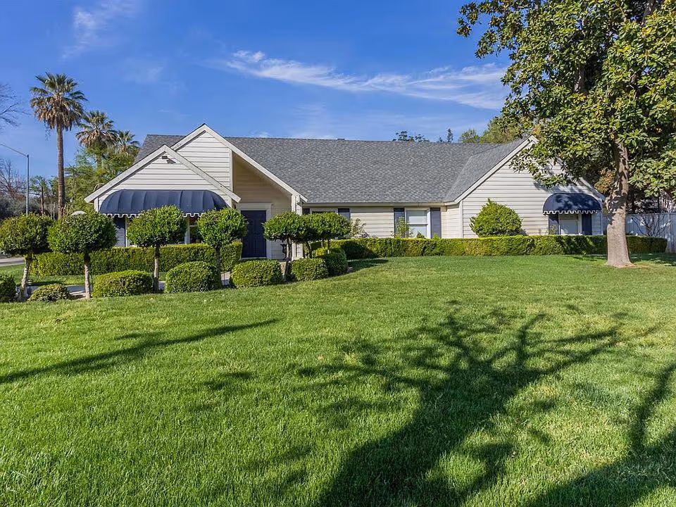 Single-story light-colored house with blue awnings, neatly trimmed shrubs and a large green lawn under a clear sky.