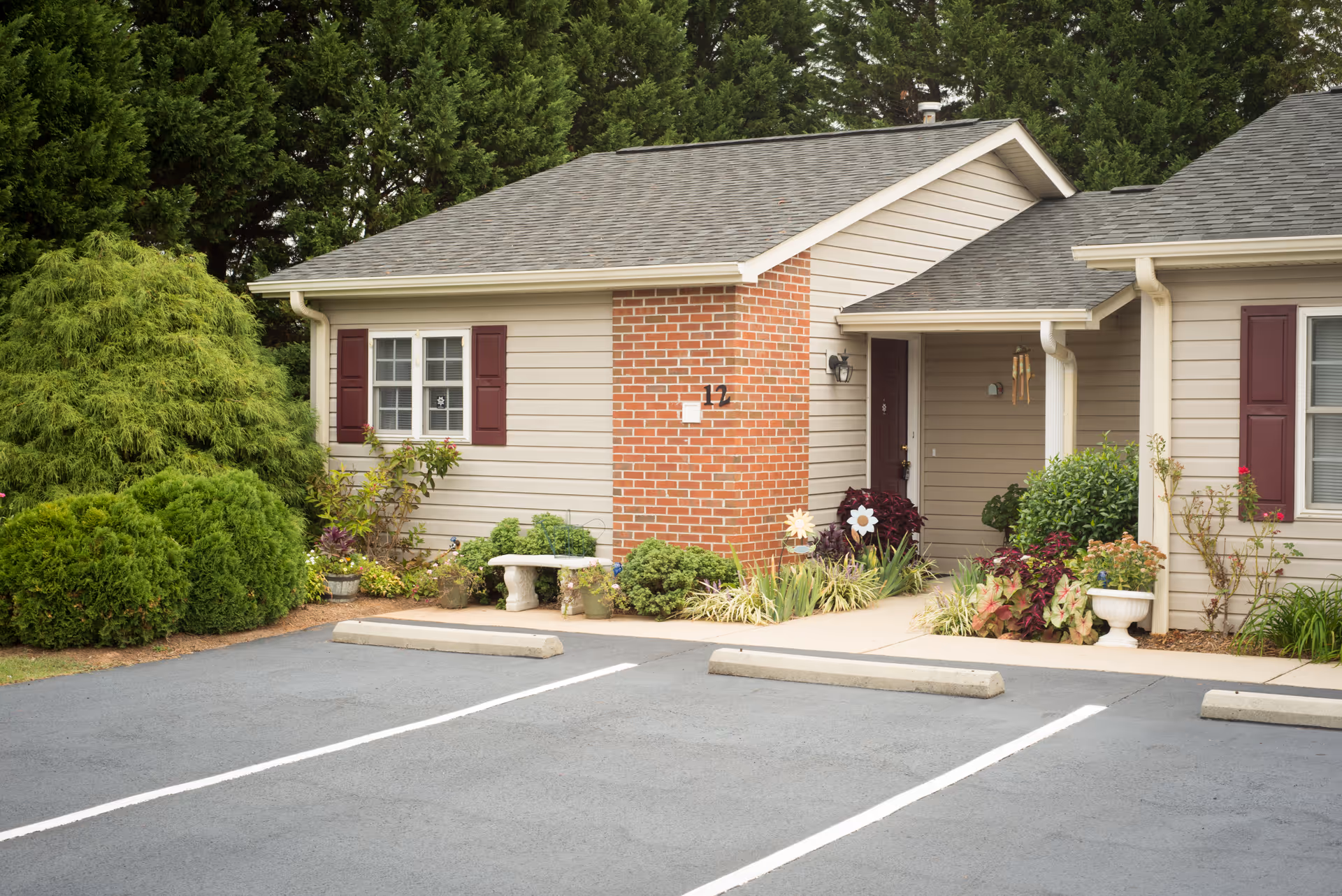 Small single-story assisted living unit with a brick entry, covered porch, landscaped plants and empty parking spaces in front.