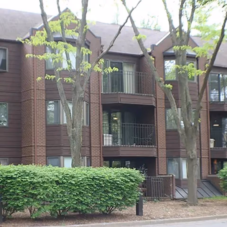 Front exterior of a multi-story brick residential building with balconies, trees, and trimmed hedges.