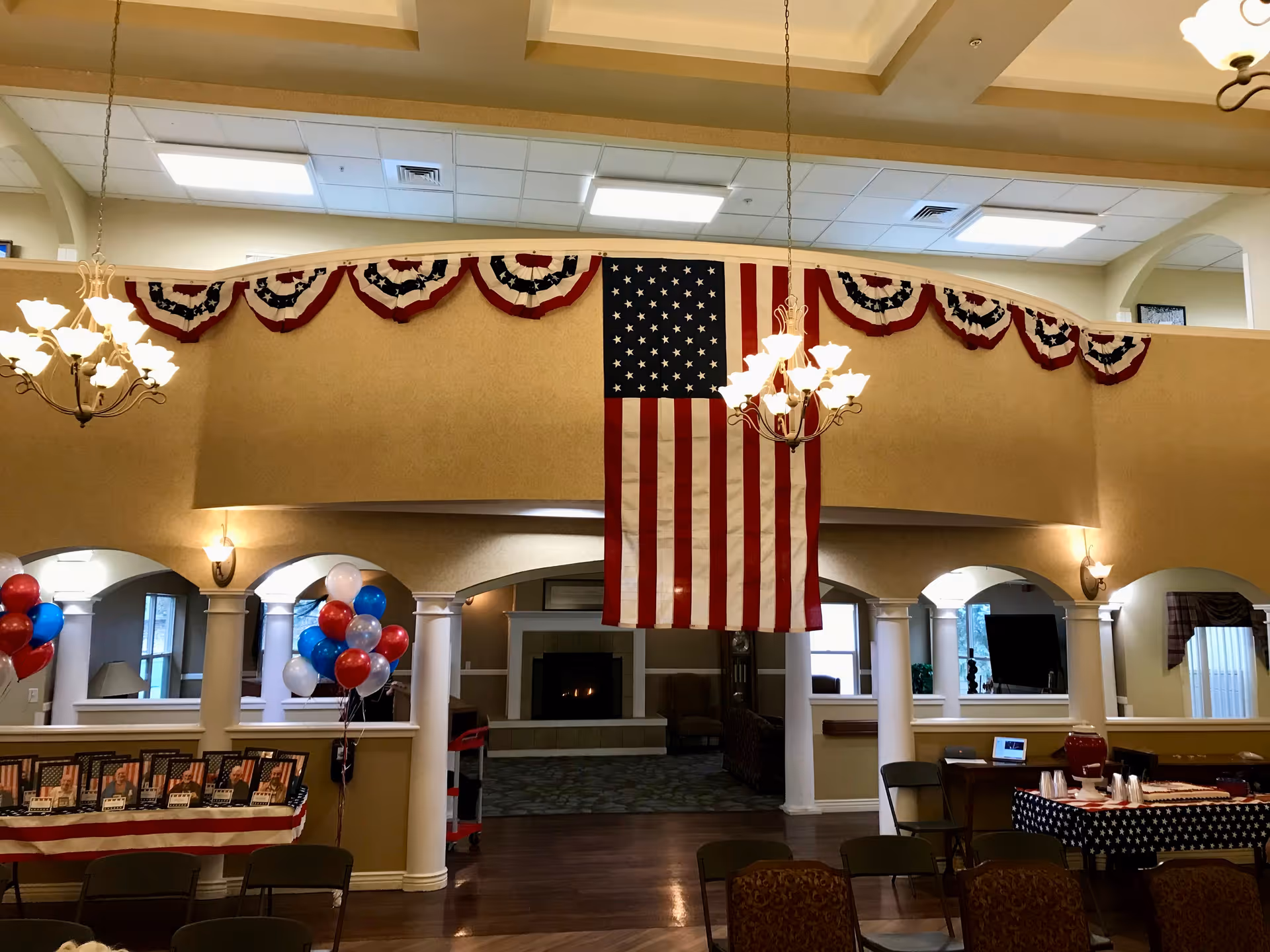 Interior view of a senior living facility decorated with patriotic red, white, and blue bunting and an American flag hanging from the balcony. There are chandeliers hanging from the ceiling, balloons in red, white, and blue colors, and tables set up with chairs. The room has columns and arches, with a fireplace visible in the background.
